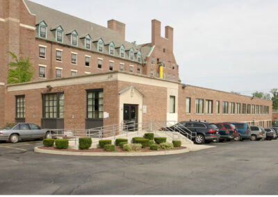 Exterior view of a large, multi-story red-brick building with teal trim and an accessible entrance ramp; several cars are parked in the foreground on an overcast day.