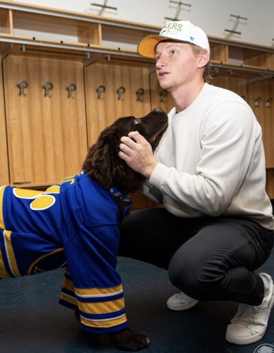 A Sabres player wearing a white cap and cream sweatshirt kneels down in the locker room to gently pet Mac, a brown Newfoundland puppy wearing a classic blue-and-gold Sabres jersey, who is gazing up adoringly.