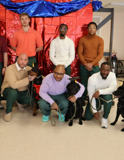 Group photo of nine men (five in green prison uniforms, four staff/trainers) with five service dogs in training (black Labs and a Doberman), posing in front of a red metallic backdrop with blue swag for a graduation or completion ceremony.