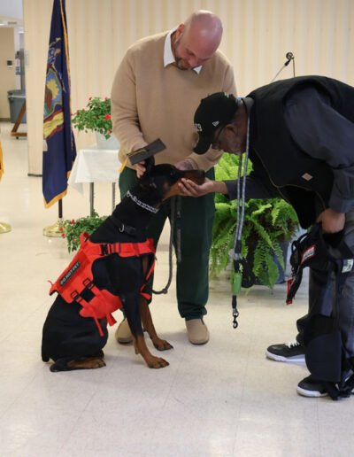 Three men indoors: one in a green prison uniform stands between two staff members; a black service dog in training wearing a vest stands calmly in front of them, with American and New York state flags in the background.