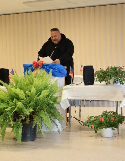 A man in a black hoodie stands at a podium reading from papers during a ceremony; the podium is decorated with American flags, red ribbons, and potted plants; a screen to the right displays a blue star with the name “GOOSE” and a photo of a dog.