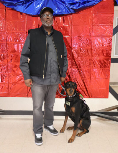 A man in a black Carhartt vest, gray shirt, and cap stands proudly holding the leash of a black-and-tan Doberman service dog in training, both facing the camera in front of a festive red metallic backdrop.