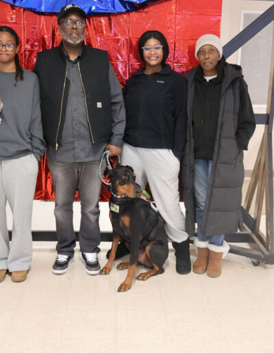 Four people (three women and one man) pose in front of a red metallic backdrop with blue swag; the man holds the leash of a black-and-tan Doberman service dog in training sitting attentively in front.