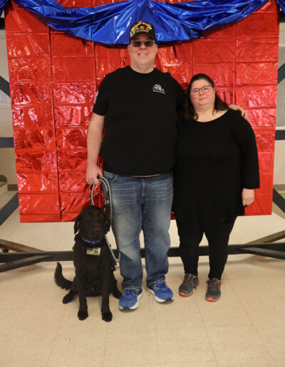 A man wearing a black t-shirt and veteran cap stands with his arm around a woman in a black dress, both smiling; a young black Labrador service dog in training sits calmly in front of them against a red metallic backdrop with blue draping.