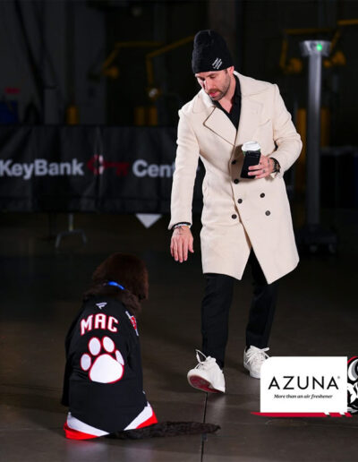 A Sabres player in a cream coat and black beanie holds a coffee cup and phone while Mac, a brown Newfoundland puppy wearing a “MAC” Sabres jersey with a large white paw print, sits perfectly in front of him on the rink floor. Azuna logo in corner.