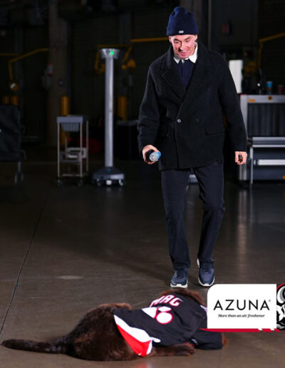 A Buffalo Sabres player in a dark peacoat and beanie smiles while holding a can and looking down at Mac, a brown Newfoundland puppy wearing a black “Girgensons 28” Sabres jersey, lying playfully on the ice rink floor. Azuna logo in corner.