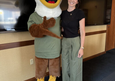A smiling woman in a black top and green pants stands next to the WNYHeroes eagle mascot who is pointing at the camera. They are indoors in front of large screens.